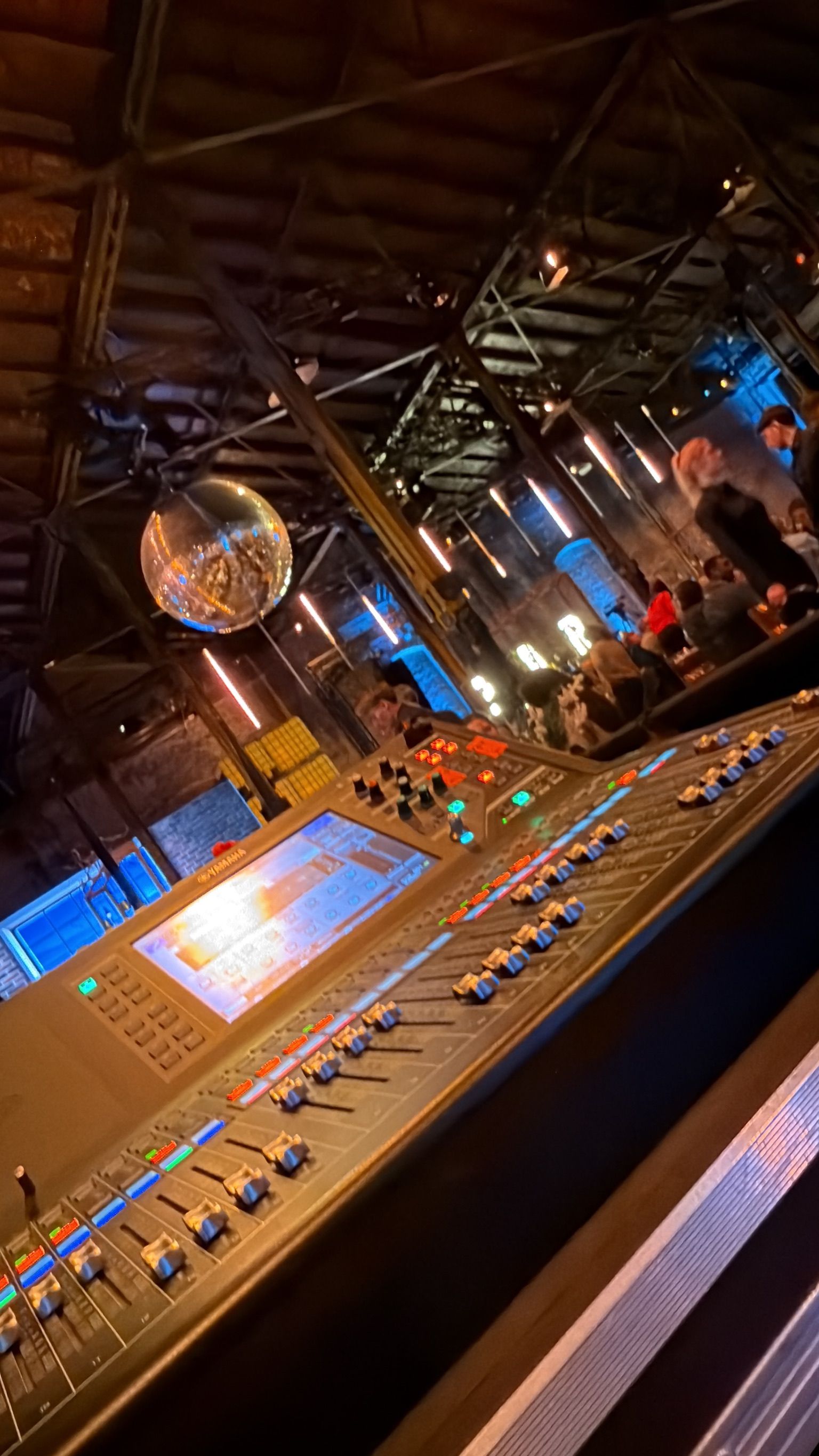 Tonmischpult in einem schwach beleuchteten Club mit Discokugel und Menschen im Hintergrund.