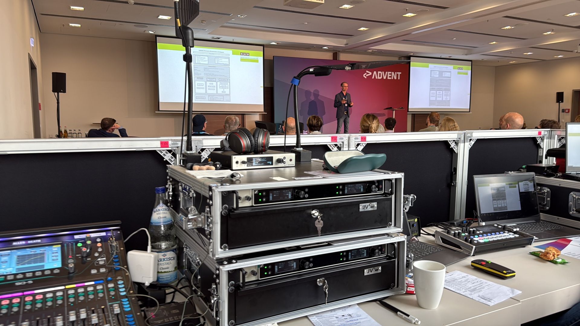 Konferenzbühnenaufbau mit Moderator, Bildschirmen, Tonanlage und Publikum.