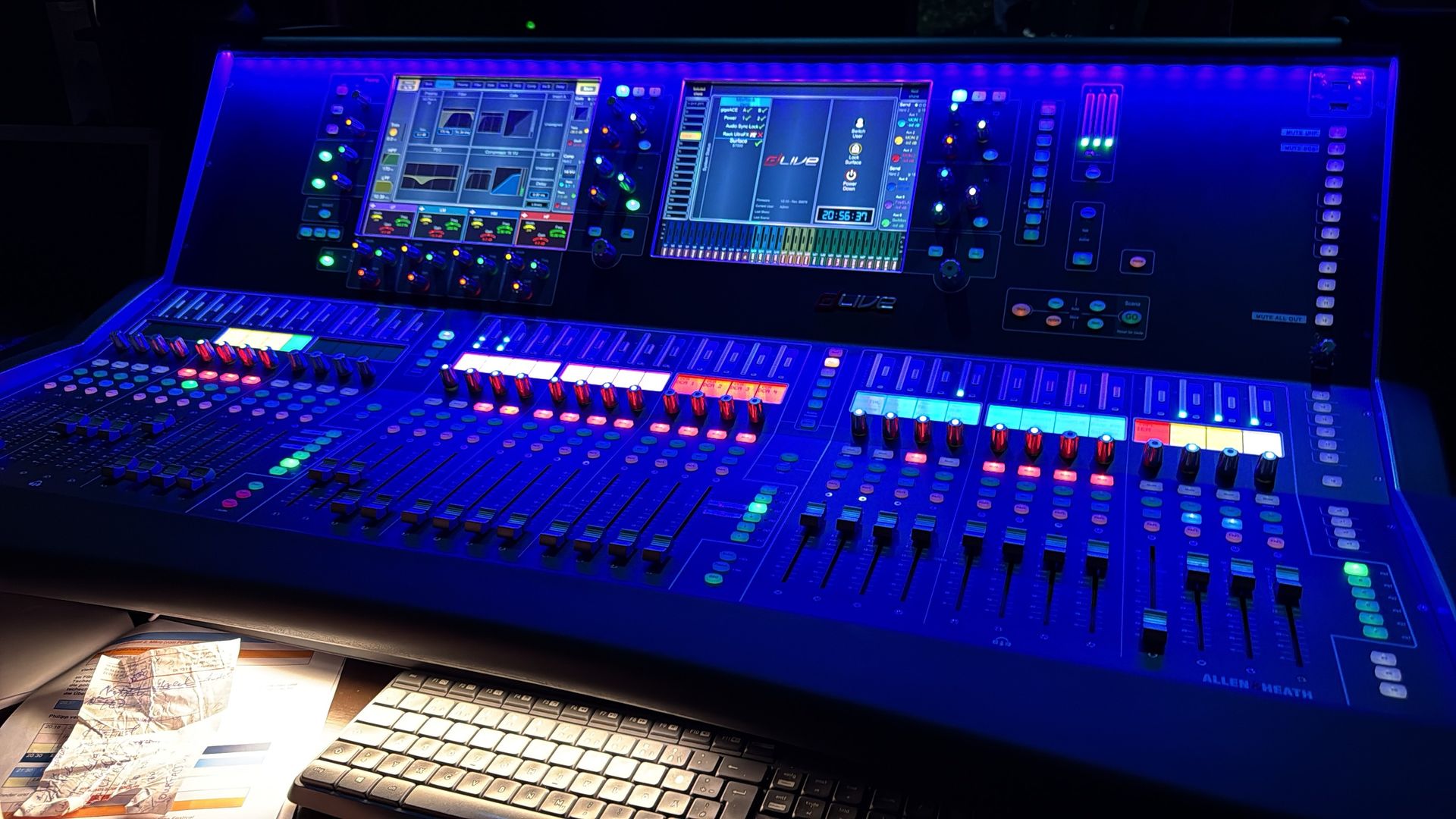Ein großes, blau beleuchtetes Audiomischpult in einem dunklen Raum mit mehreren Bildschirmen und Fadern.
