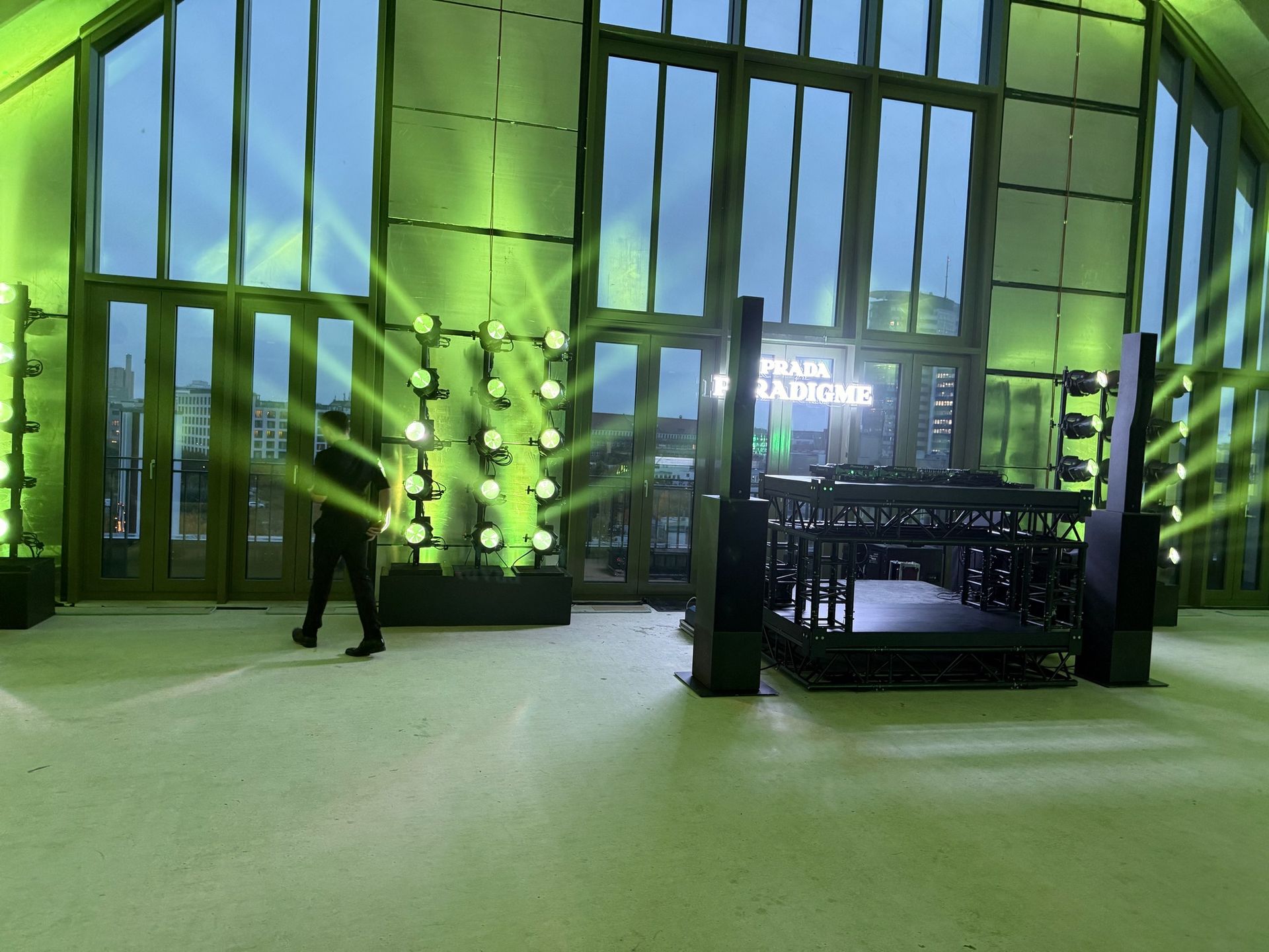 Veranstaltungsraum mit grüner Beleuchtung, Bühne, Stühlen und Logo; eine Person geht durch die Tür.