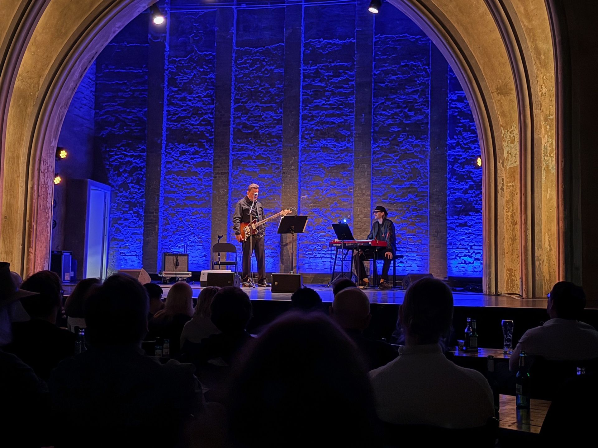 Zwei Musiker treten auf einer mit blauem Licht beleuchteten Bühne vor Publikum auf.