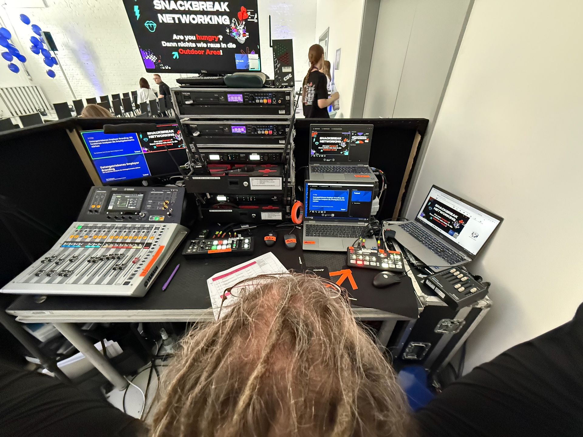 Eine Person mit langen Haaren arbeitet an einem Medien-/Technik-Setup mit Mischpulten, Computern, Bildschirmen und Geräteracks auf einer Konferenz.