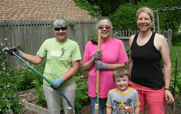 Three women and a child, holding gardening tools, stand in front of the Heritage House colonial four-square garden.